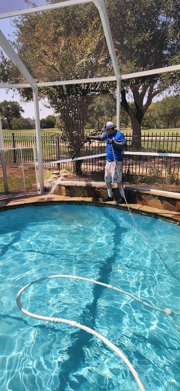 A person in blue coveralls uses a pressure washer to clean a wooden deck outside a house.