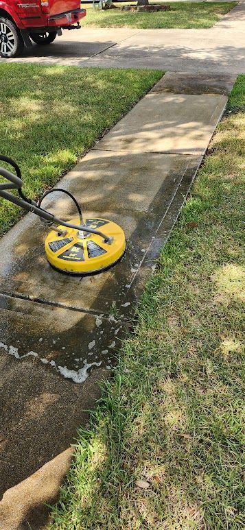 Yellow pavement cleaner on a wet sidewalk beside a grassy lawn and driveway, with a red truck in the background