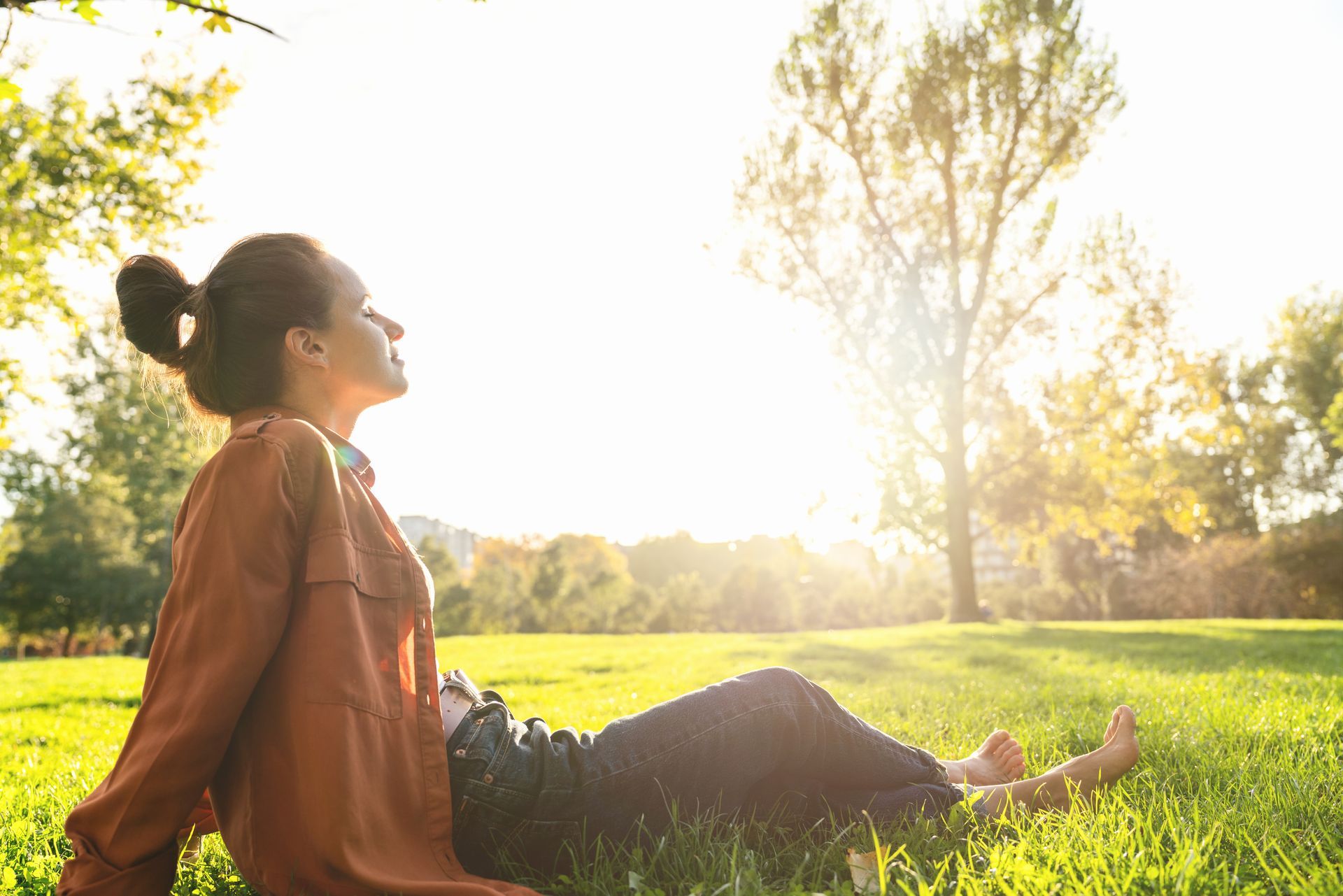 Frau sitzt im Gras, blickt in die Sonne, entspannter Gesichtsausdruck. Goldenes Sonnenlicht und grüne Bäume im Hintergrund.