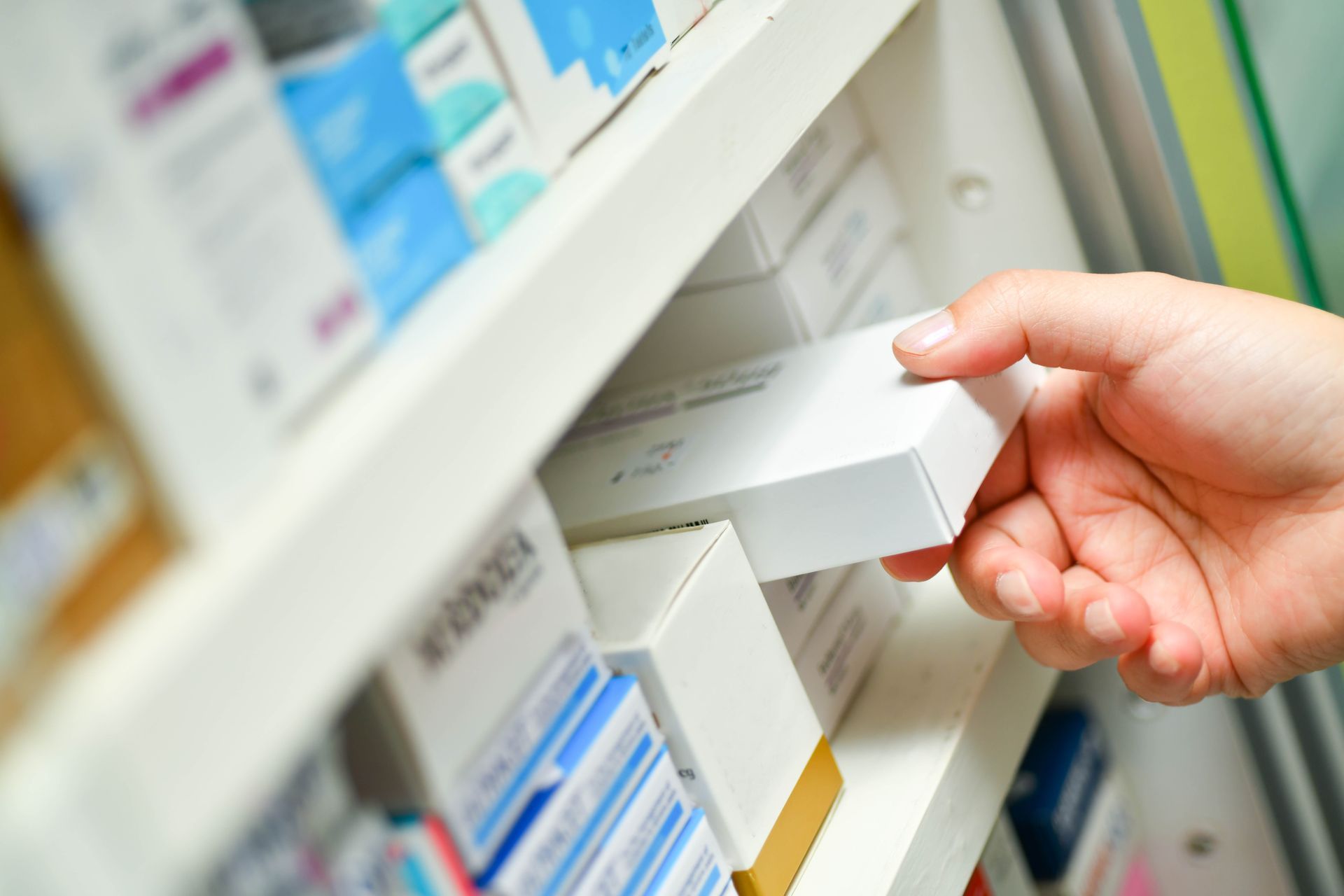 Hand greift nach einer weißen Medikamentenschachtel auf einem Apothekenregal, das mit verschiedenen Medikamentenschachteln gefüllt ist.