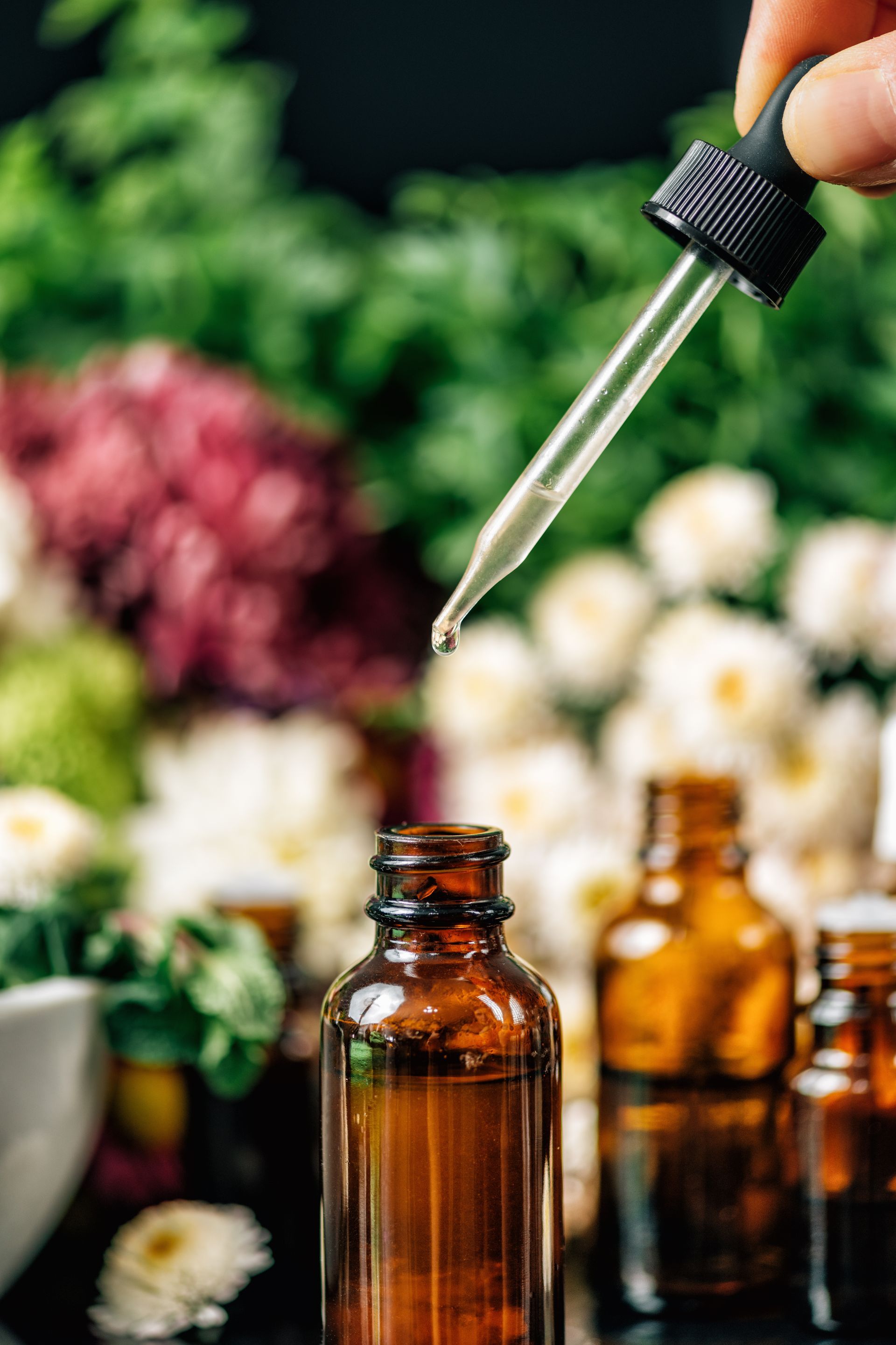 Hand hält eine Pipette über einer braunen Flasche; Blumen im Hintergrund.