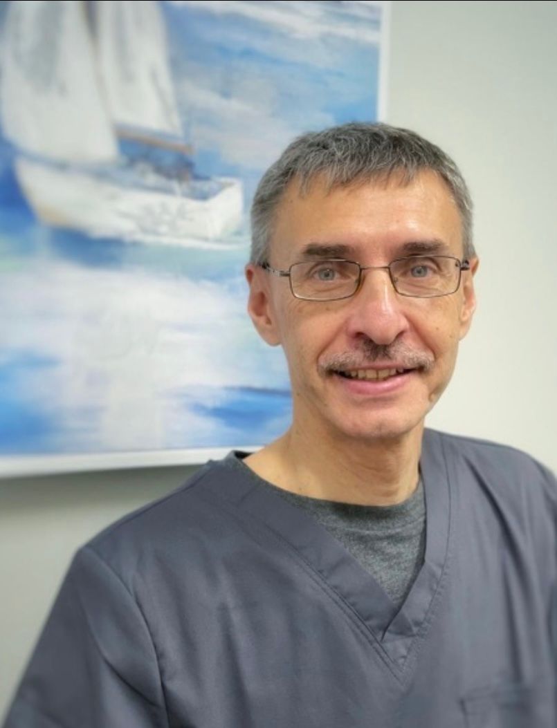 A man, Igor Krivoshey, wearing glasses is smiling in front of a painting of a sailboat