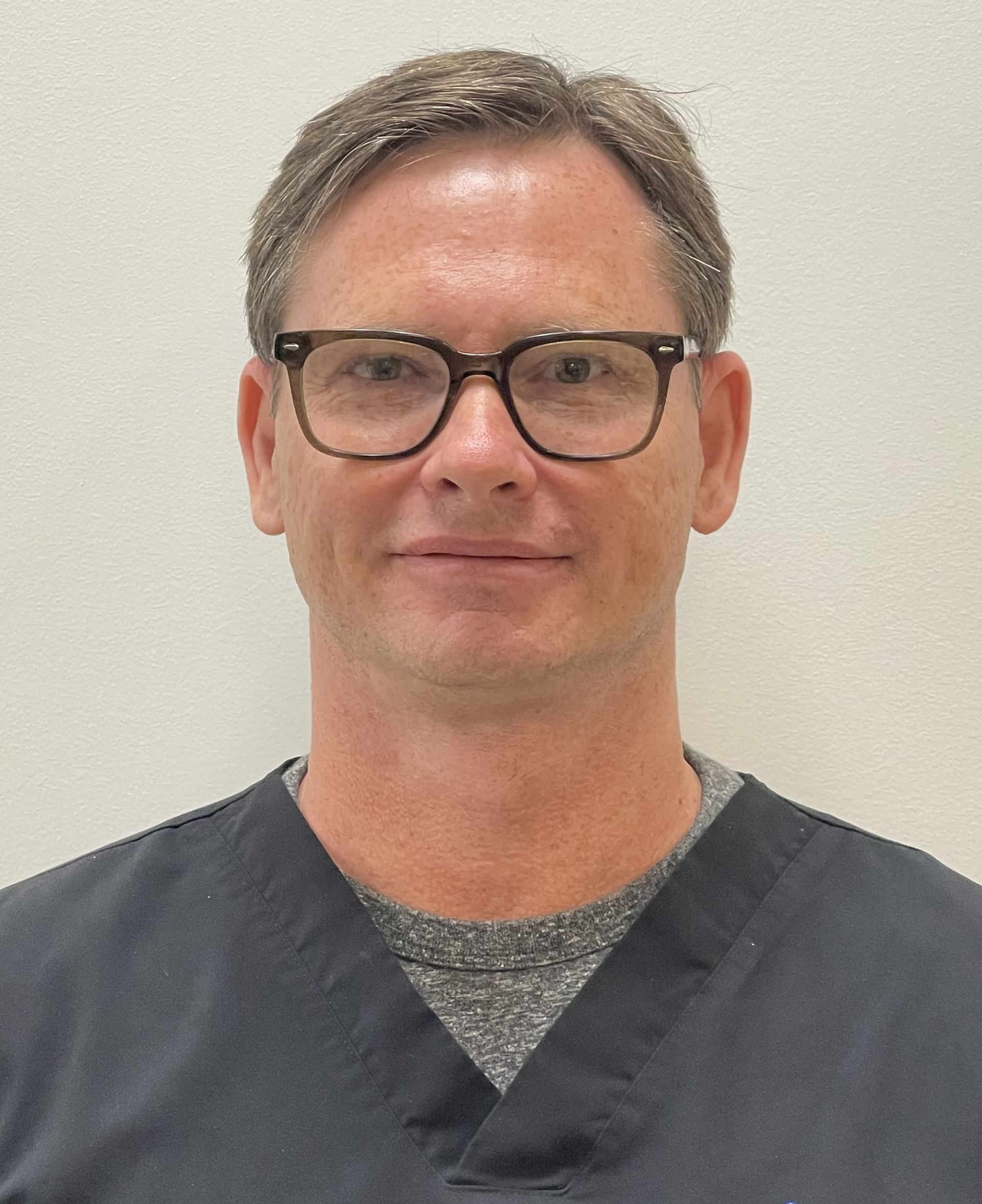 A man, Dustin Caudill, wearing glasses and a scrub top is smiling for the camera.