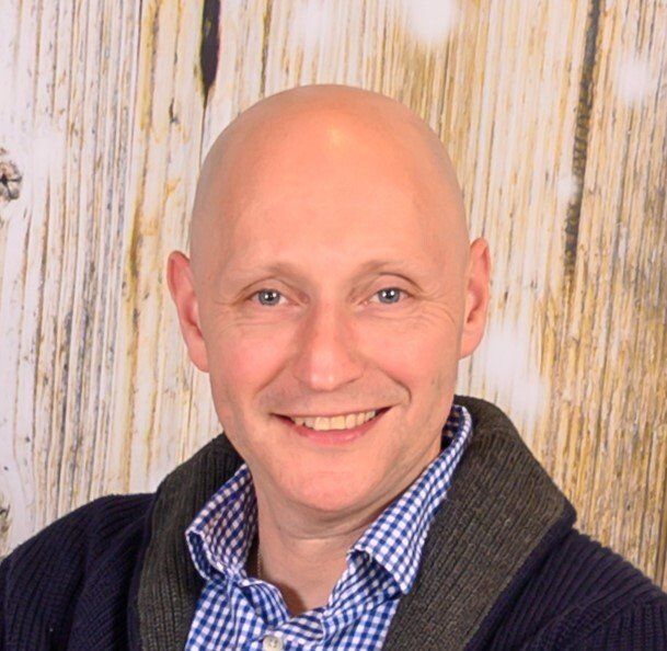 A bald man, Lev Volosskiy, wearing a sweater and a plaid shirt is smiling in front of a wooden wall.