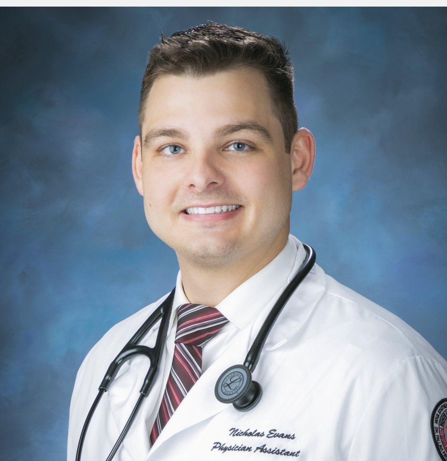 A man, Nicholas Evans, wearing a white coat and tie with a stethoscope around his neck