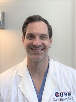 A man, Dustin Caudill, wearing glasses and a scrub top is smiling for the camera.