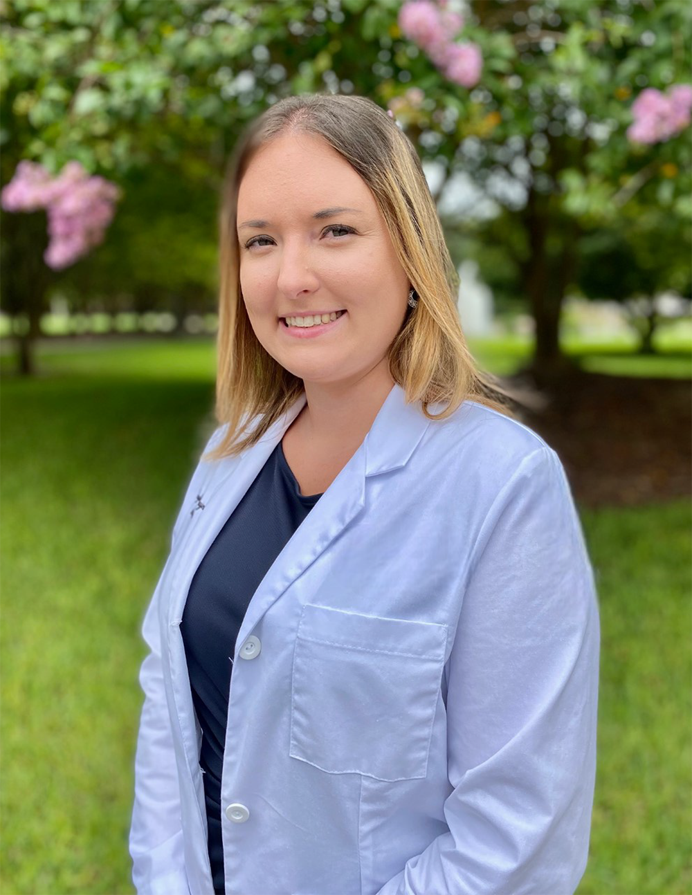 A woman, Taylor Haynes, in a white lab coat is standing in front of a tree.