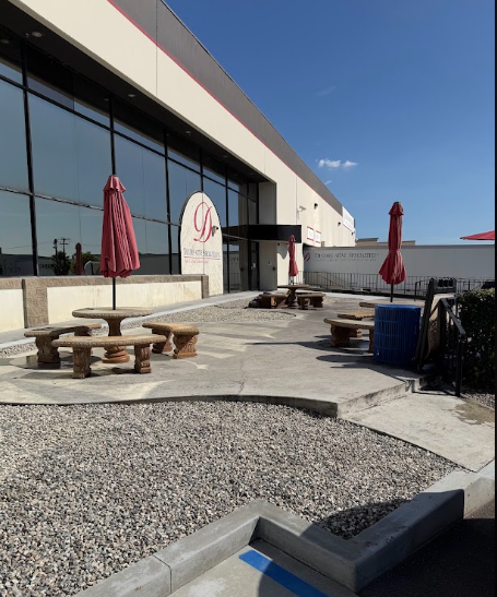 Outdoor patio with picnic tables and red umbrellas beside a glass-fronted building under a blue sky