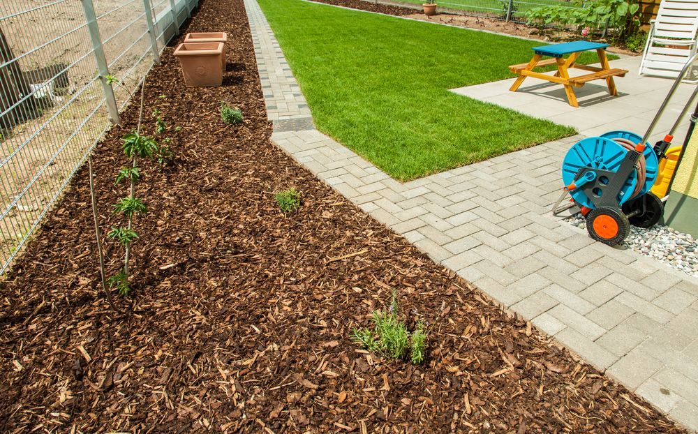 Garden with fresh mulch bed, newly planted seedlings, paved path, and lawn with picnic table and wheelbarrow.