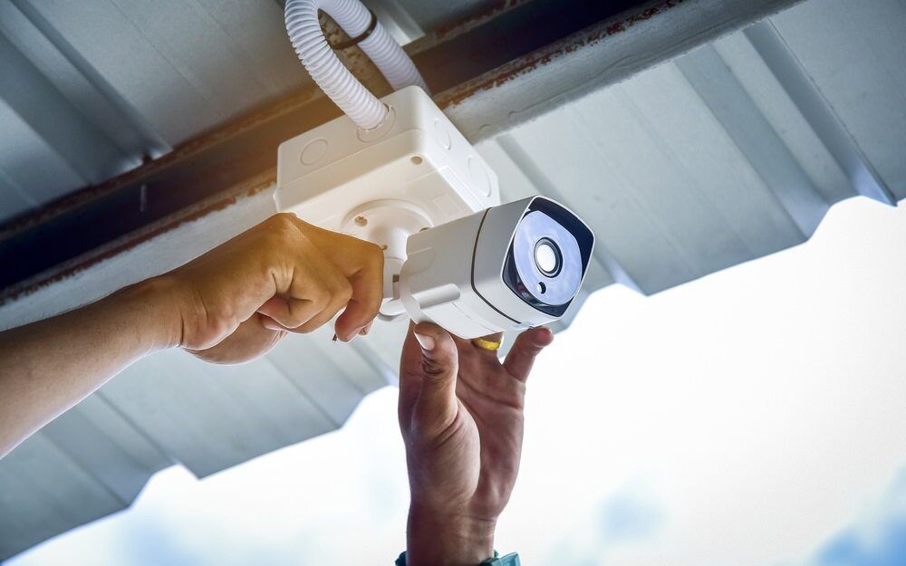 Hands Installing a White Security Camera Under a Roof — Jendata In Northward, QLD