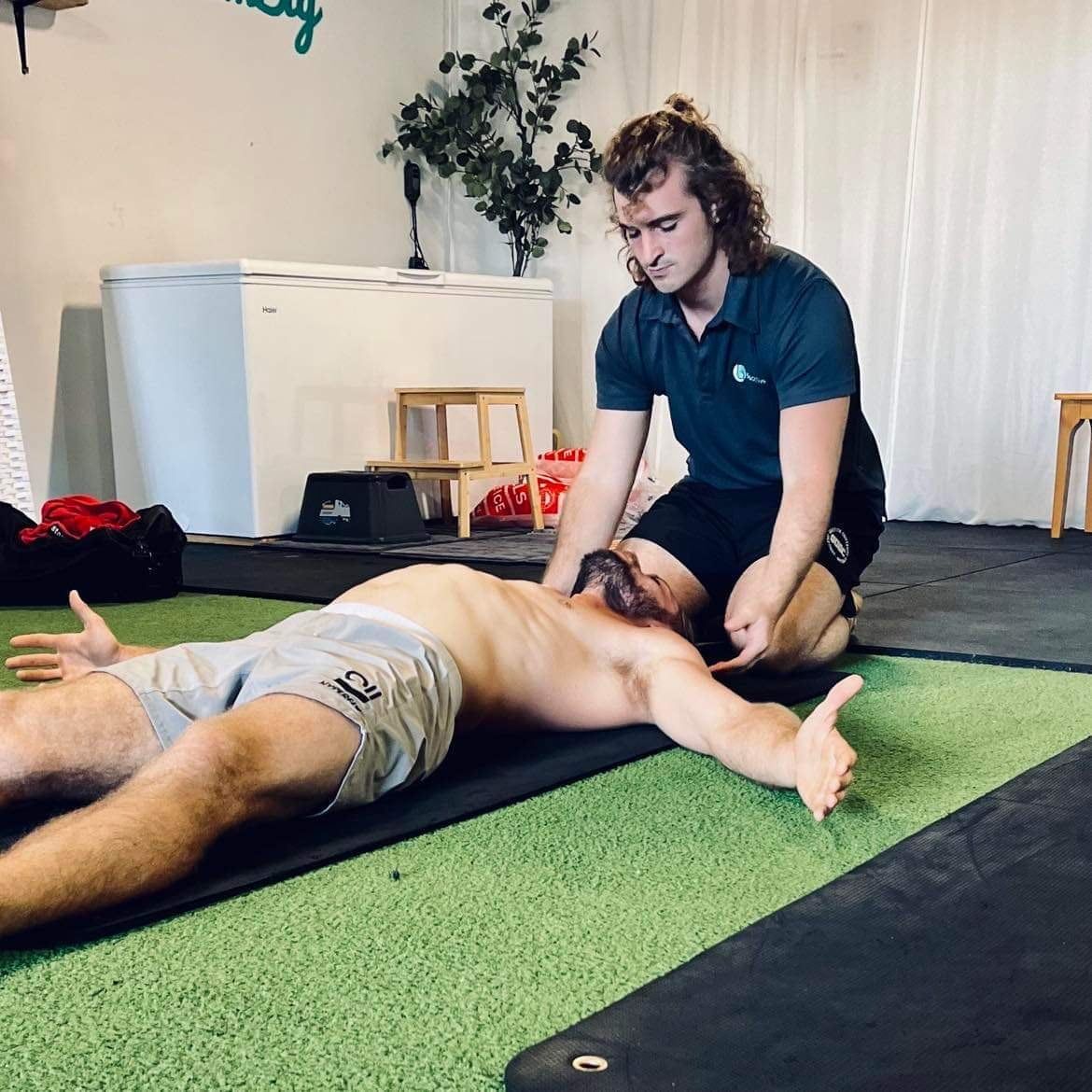 A man is laying on a yoga mat while another man kneels next to him.