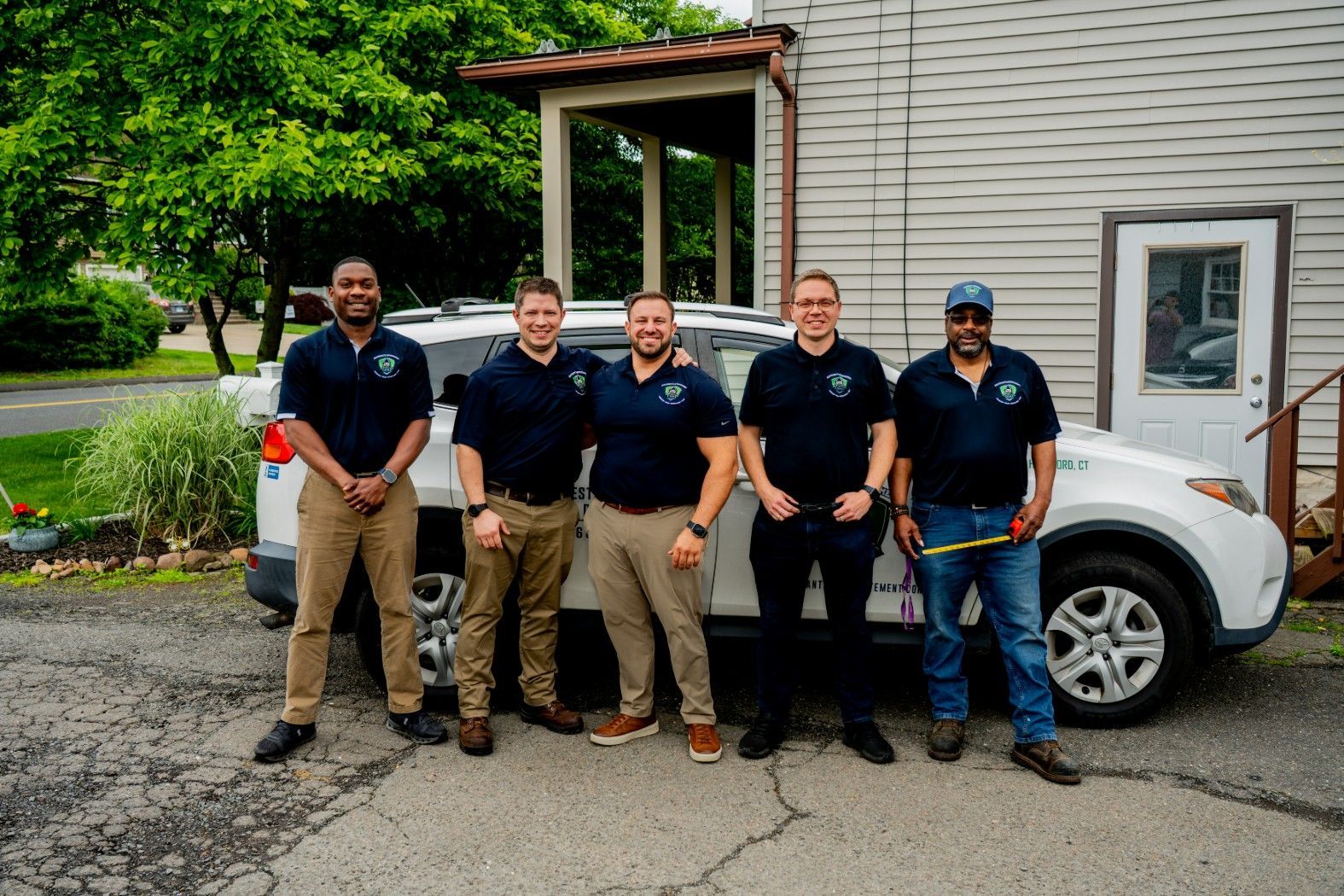 Five workers in navy polos and khaki pants stand beside a white work truck outside a building.