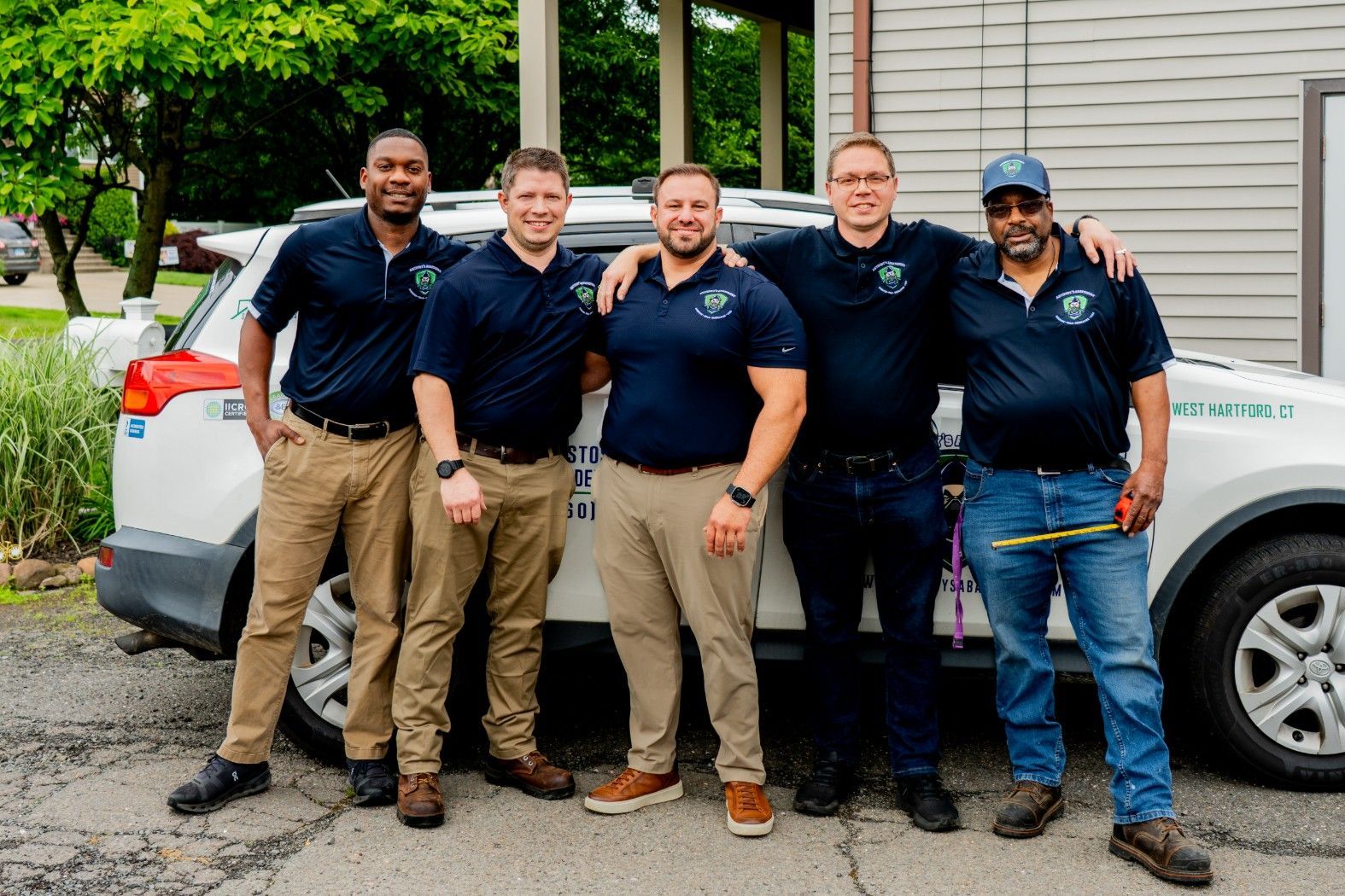 Five workers in navy shirts and khaki pants stand smiling beside a white vehicle outdoors.