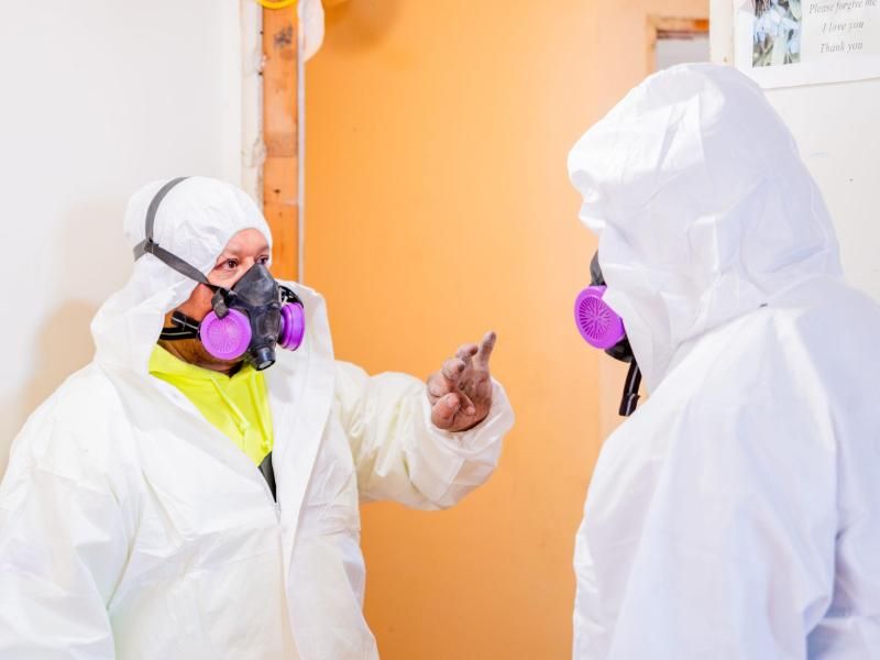 Two asbestos abatement workers in full protective gear, including white hazmat suits and purple respirators, having a discussion during an asbestos removal project.
