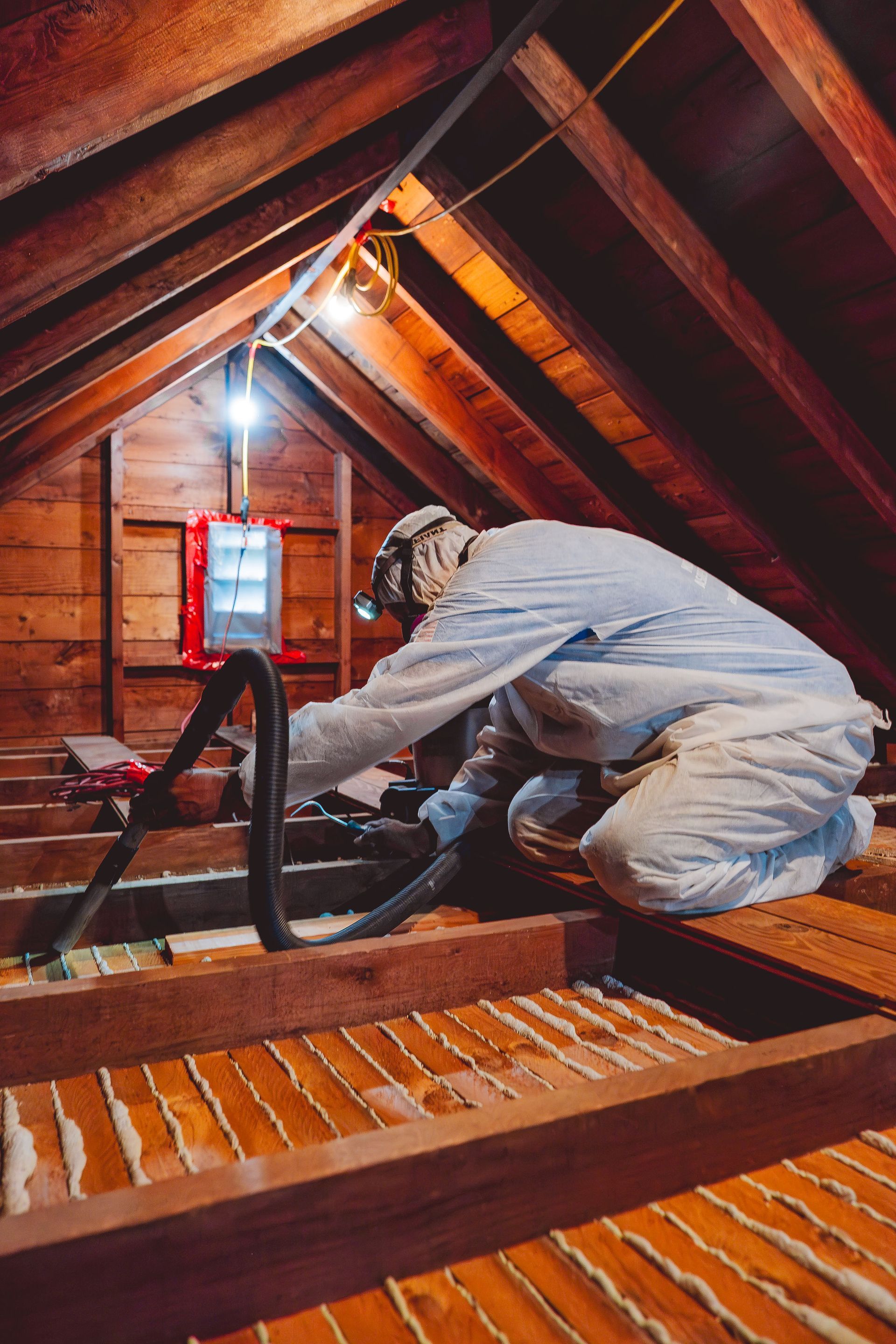 Workers in protective suits and masks removing insulation from an attic. Wooden beams and string lights are visible.