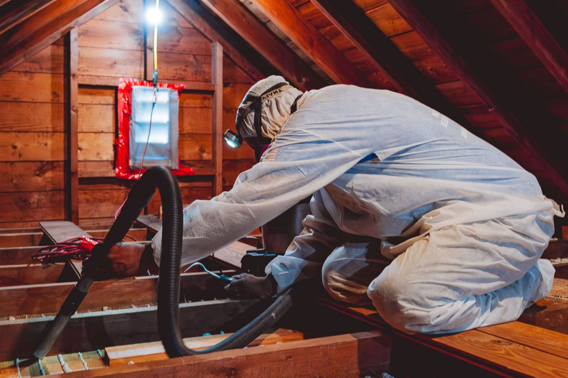 Person in protective suit vacuums attic insulation. Brown wood beams, red tool, overhead light.