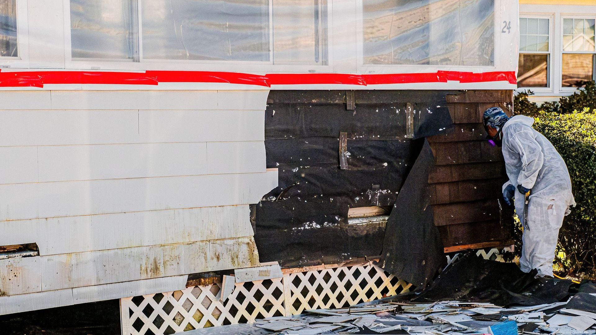 Two men in protective suits are working on removing asbestos siding.
