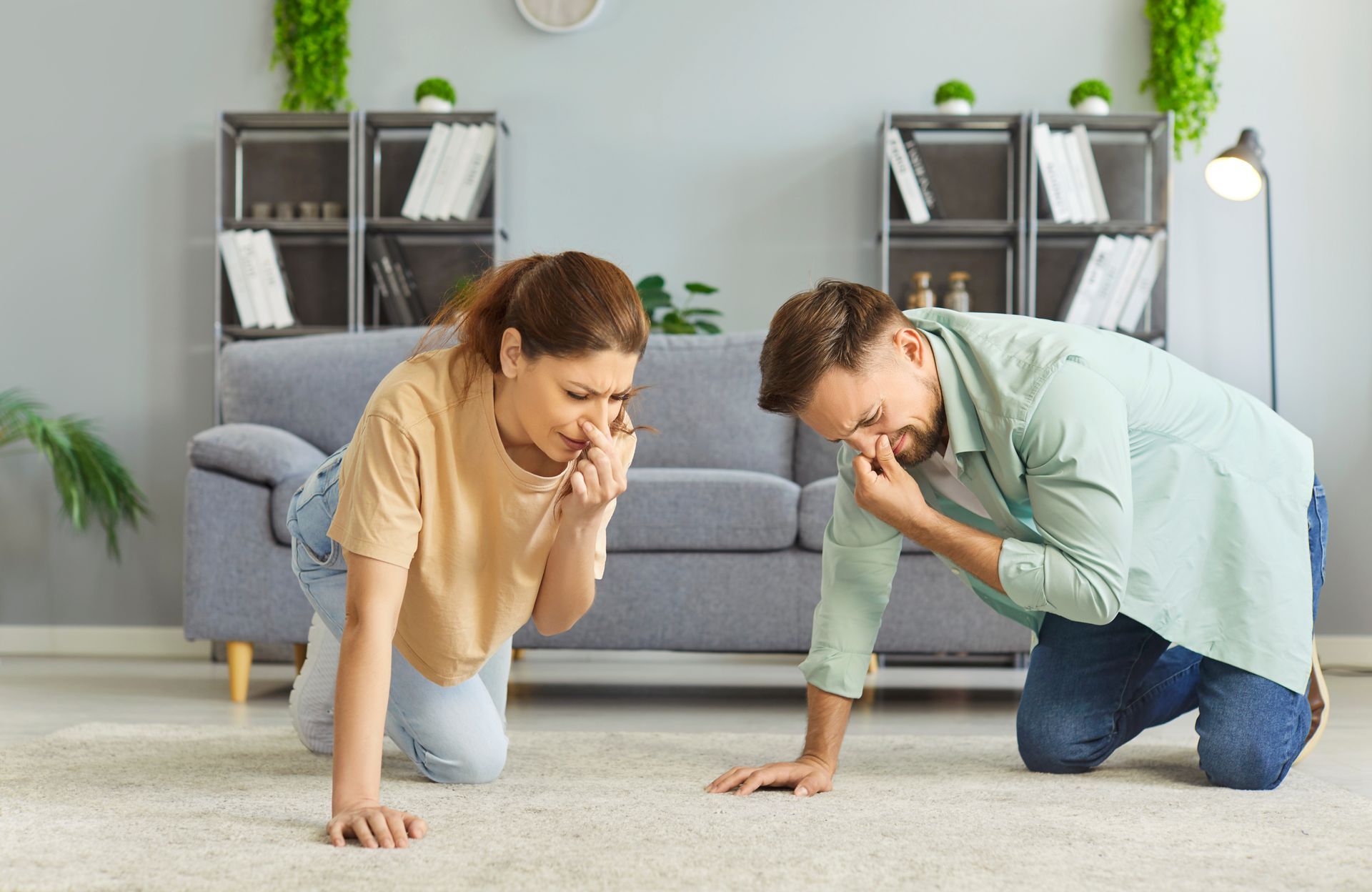 A couple kneeling on a carpet, holding their noses. They appear to be smelling something unpleasant.