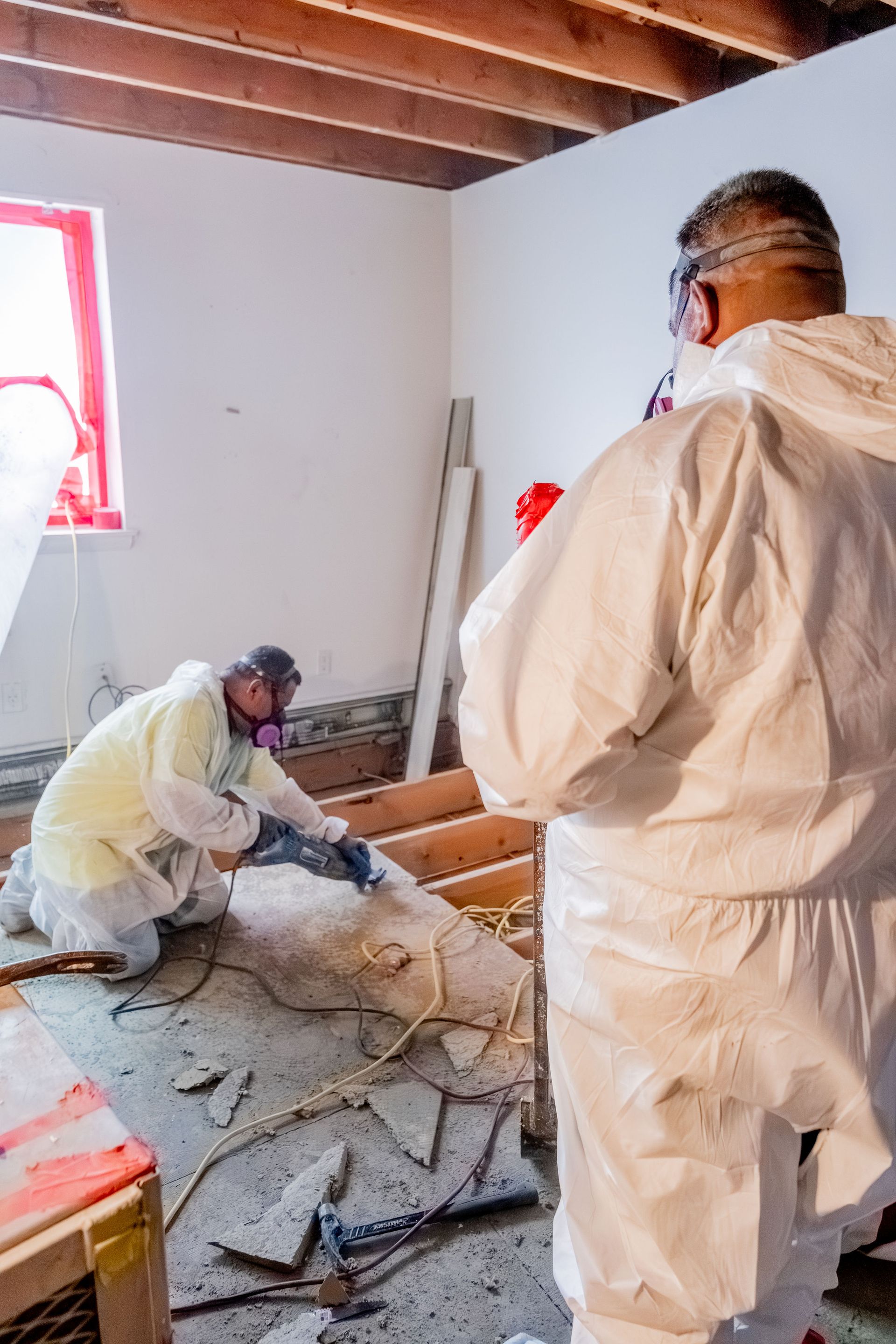 Two people in protective suits removing debris from a room; exposed wooden beams and a window are visible.