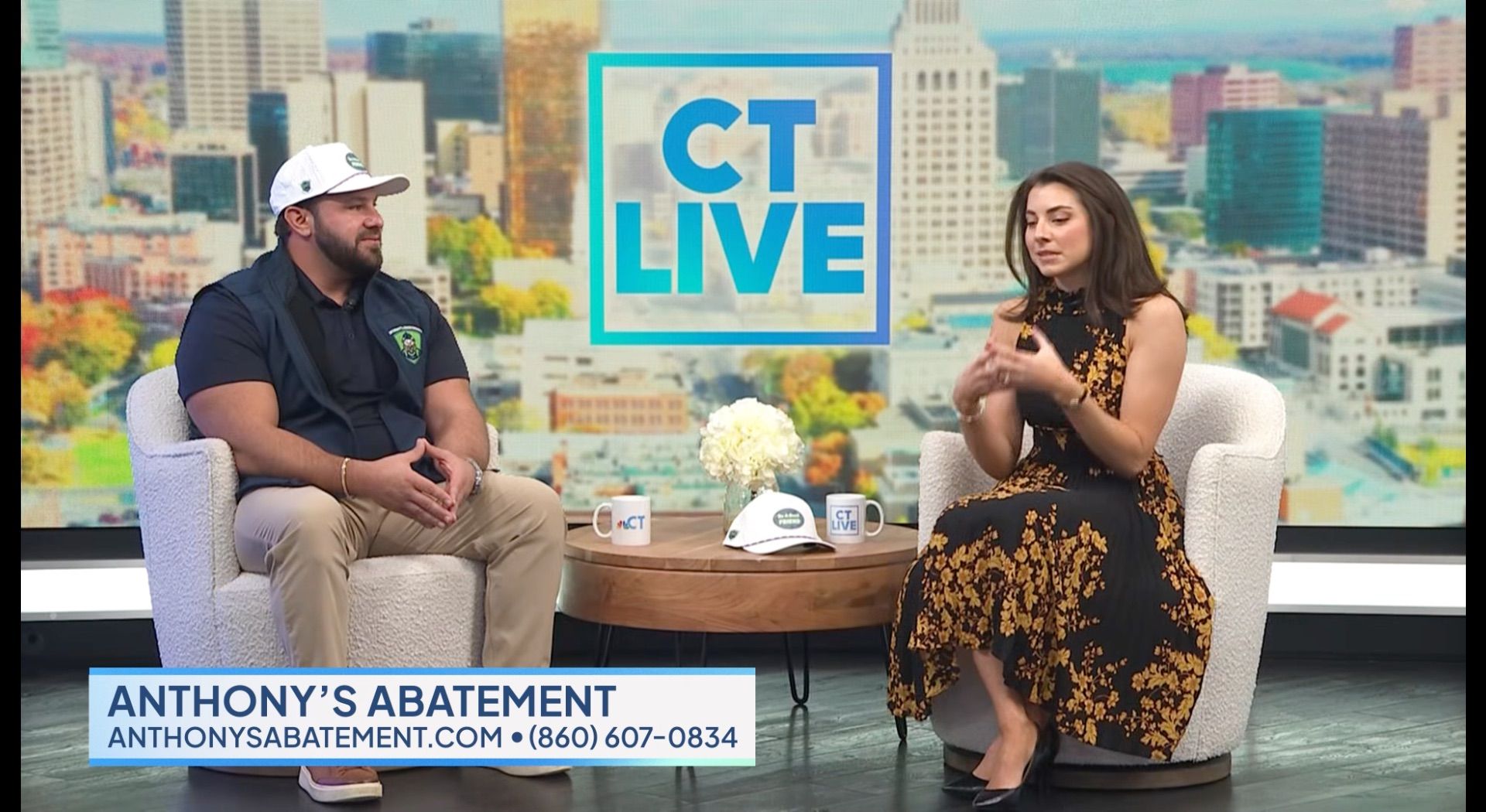 Man in work clothes and woman in dress sit on a talk show set in front of a city skyline.