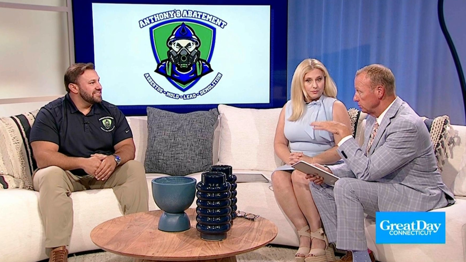 Three people on a TV set with a logo in the background. They are sitting around a coffee table, talking.