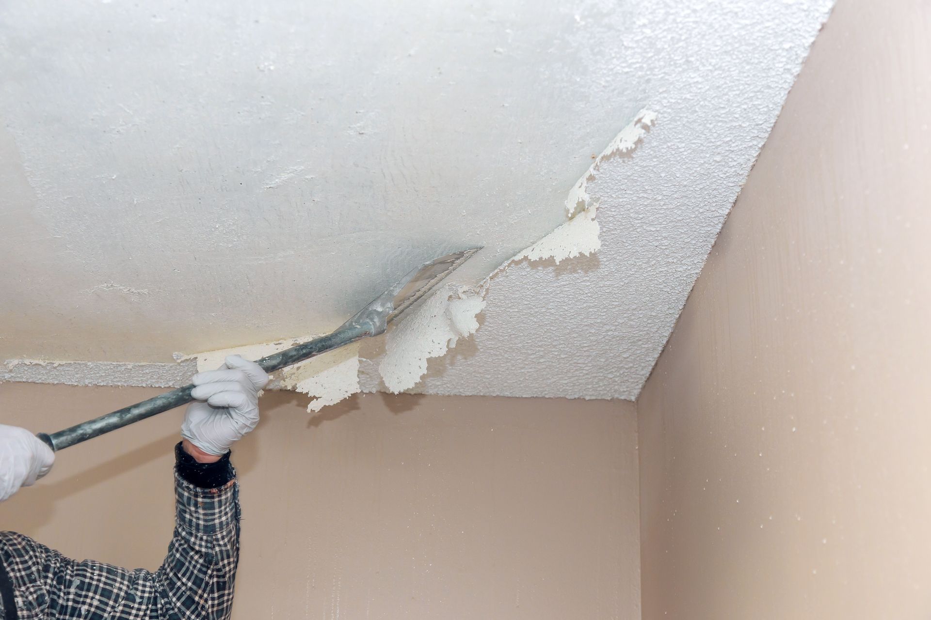 A gloved hand uses a long-handled metal scraper to remove textured popcorn ceiling material from a white ceiling.
