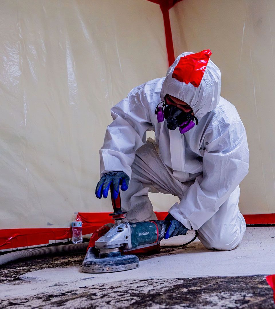Person in a white hazmat suit grinding concrete inside a containment tent, with safety mask and gloves.