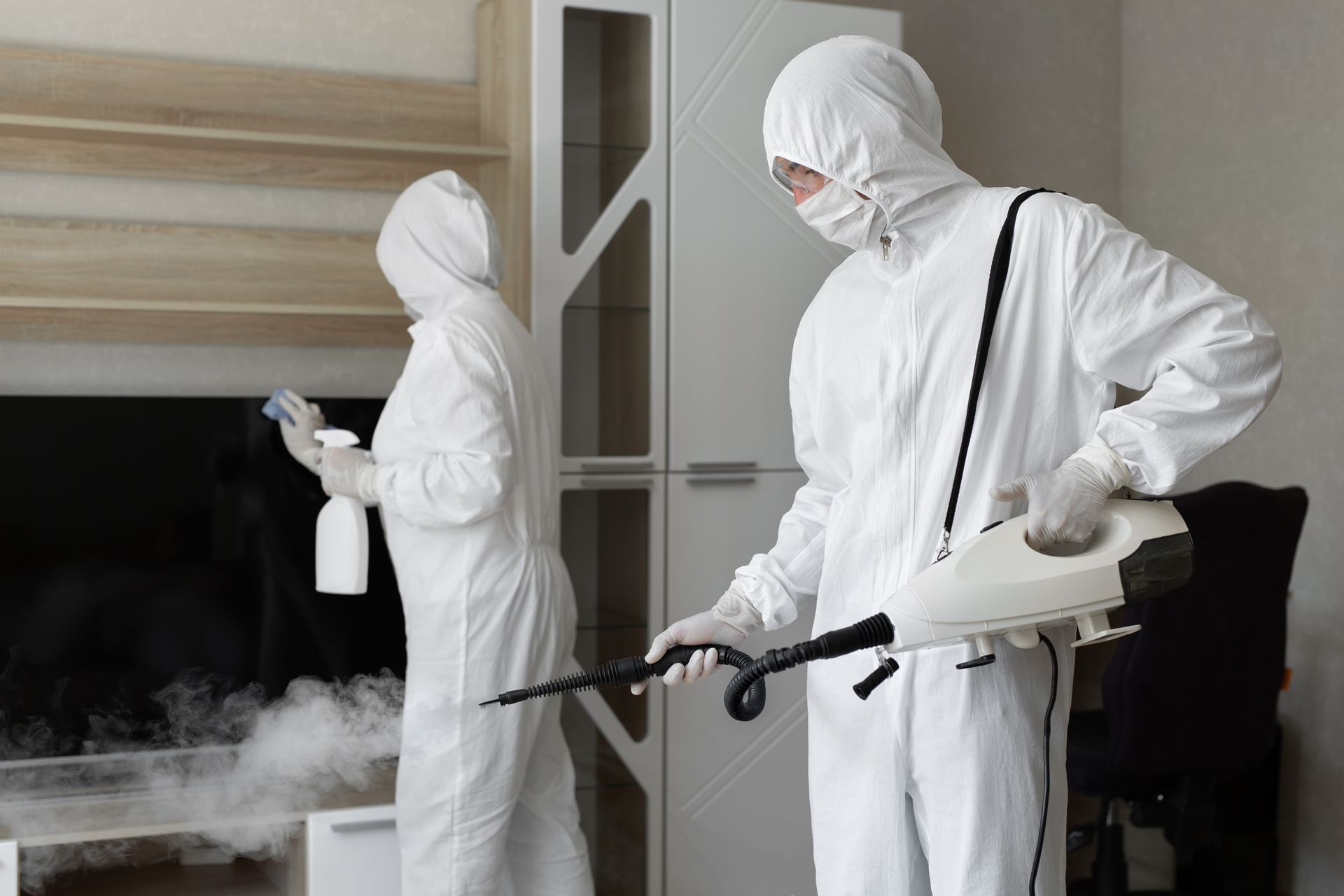 Two people in white hazmat suits sanitizing a room with sprayers and cleaning supplies.