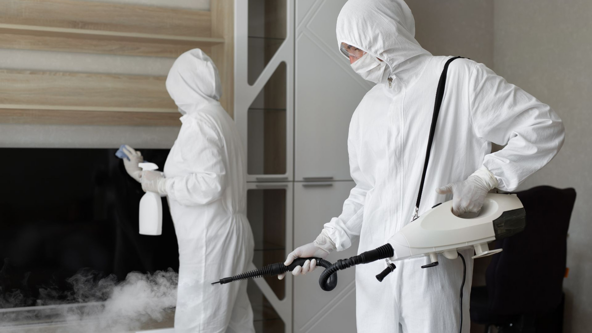Two people in white hazmat suits disinfecting a room, one spraying and the other with a fogger.