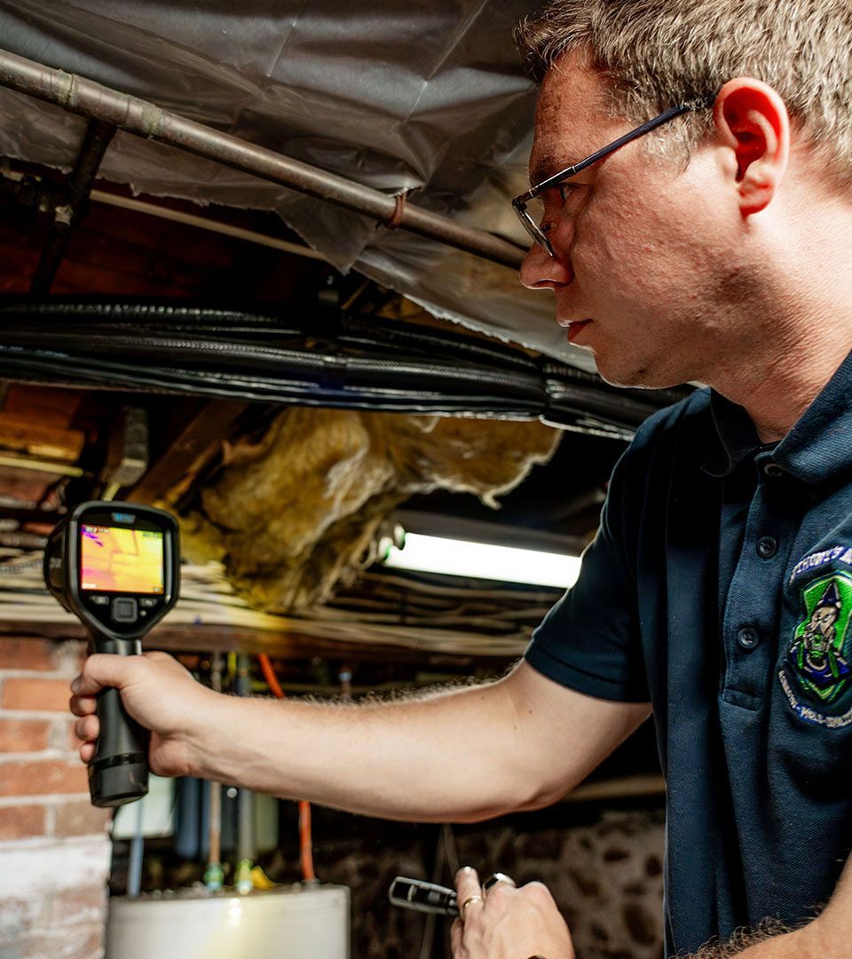 A man using a thermal camera examines plumbing in a basement.