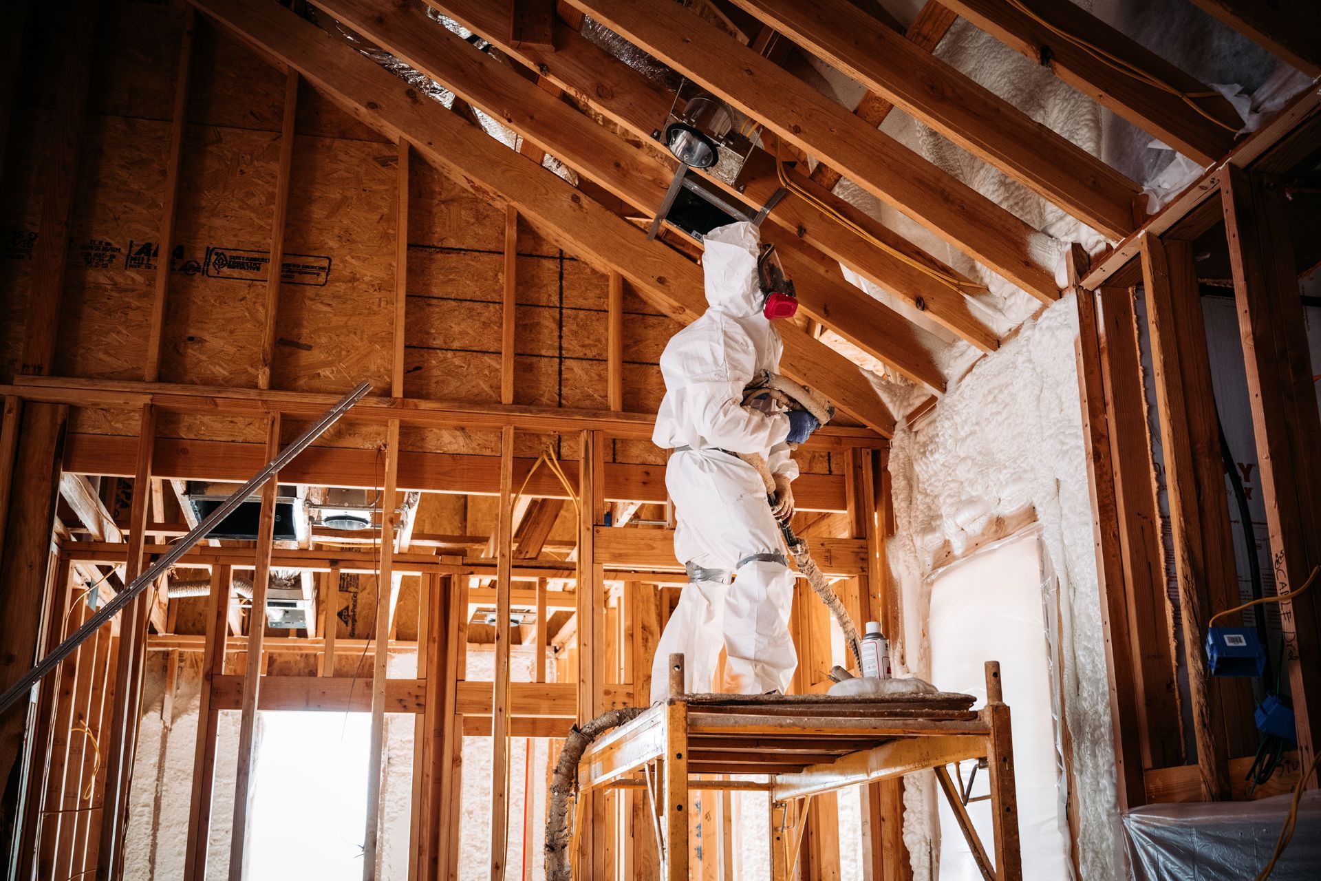 A worker in a white hazmat suit and respirator sprays foam insulation onto the rafters of an unfinished wooden house.