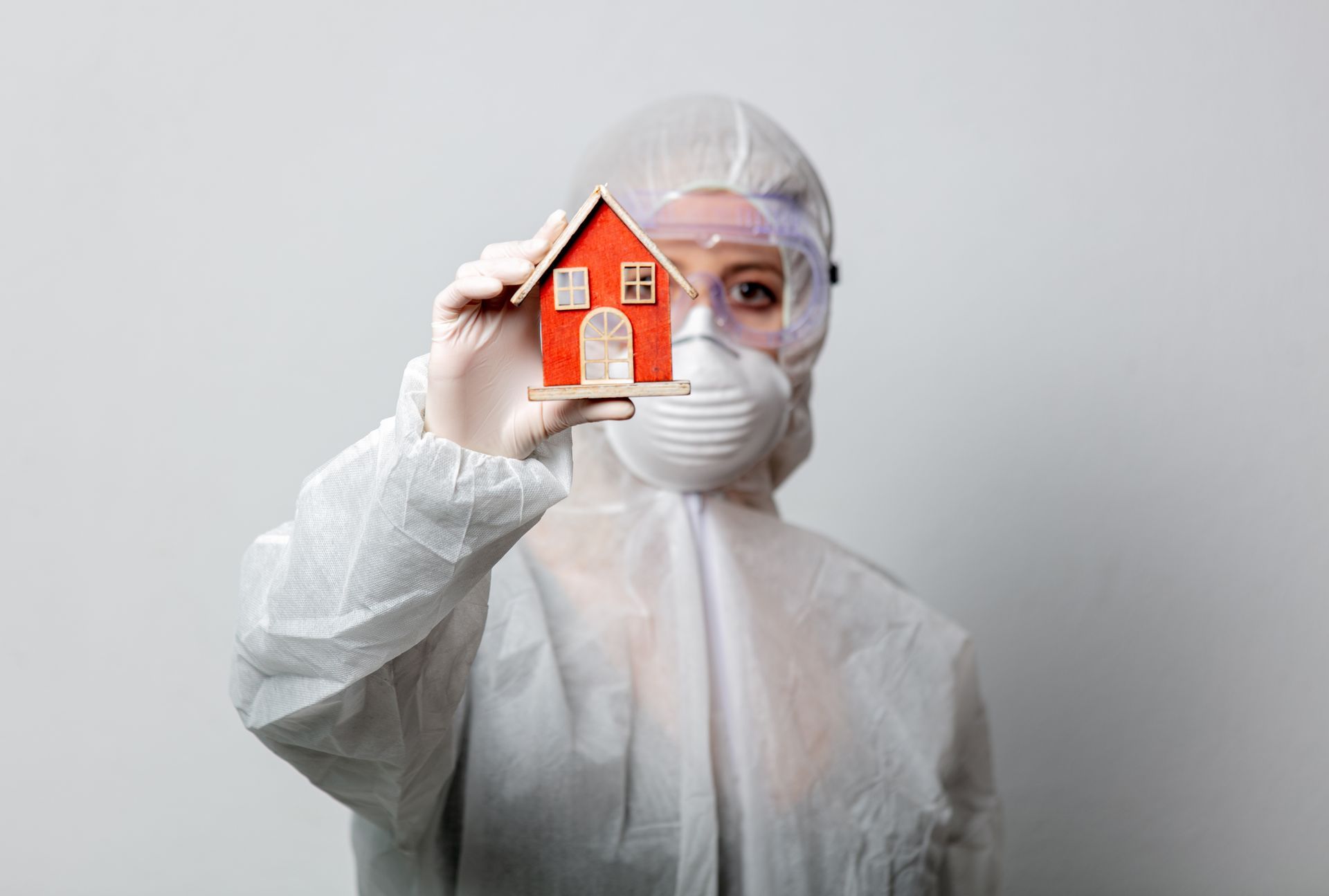 Person in protective suit holding a small red model house against a gray background
