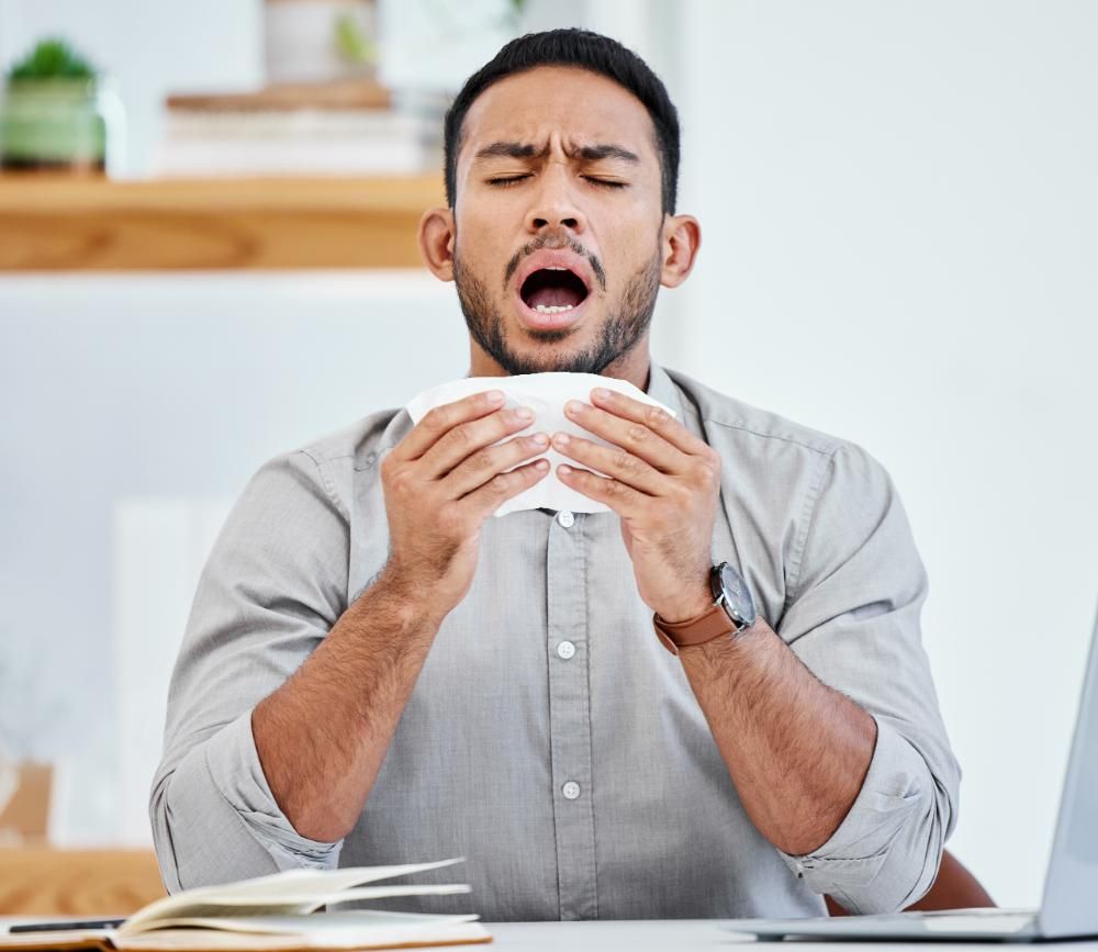 A man is sneezing into a napkin while sitting at a desk.