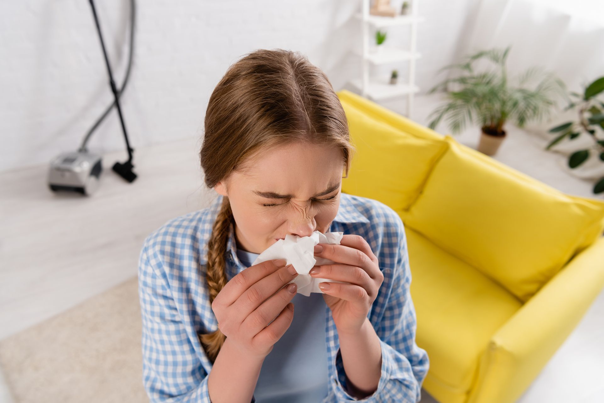 Woman sneezing into a tissue on a couch in a bright living room