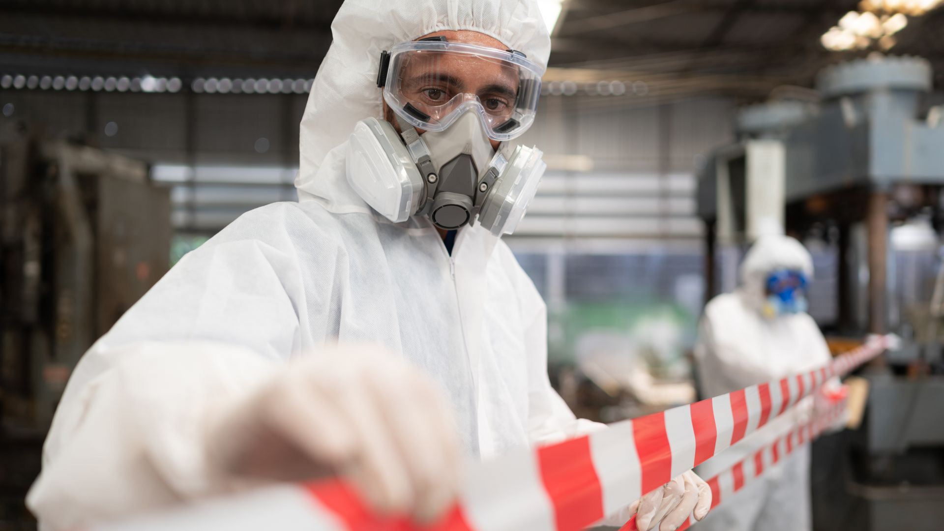 Person in hazmat suit, respirator, and goggles holds red and white barrier tape in a workshop setting.