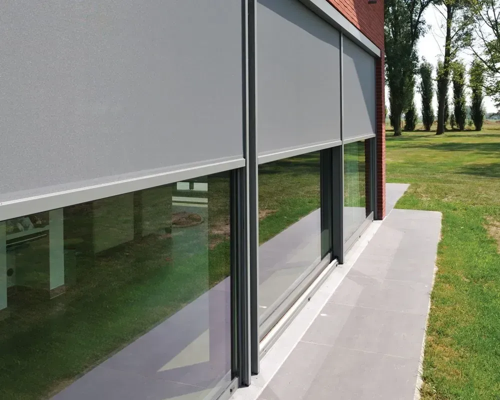 Bygningens udvendige visning med grå rullegardiner, der dækker vinduerne. Grønt græs og træer synlige.