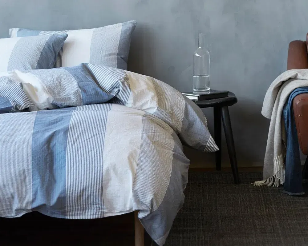 Blå og hvidstribet sengetøj på en seng, ved siden af et lille bord med en vandflaske.