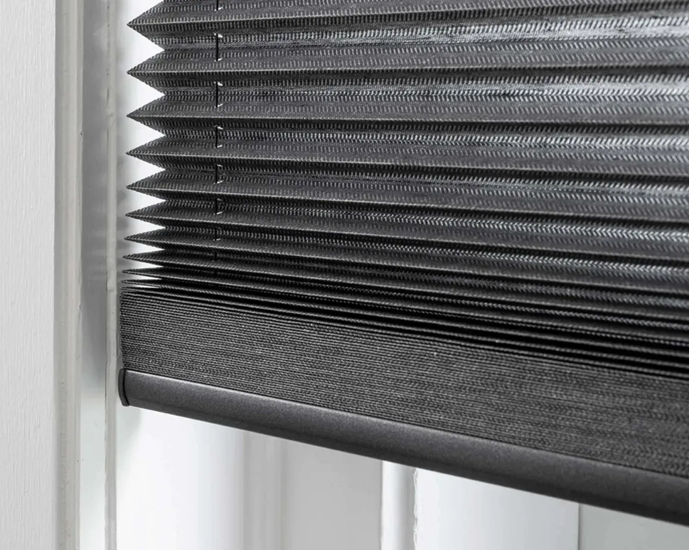 Nærbillede af en mørkegrå plisseret vinduesgardin delvist sænket, sat op mod en hvid vinduesramme.