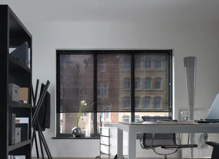 Kontorinteriør med sorte persienner, der dækker et vindue; et skrivebord, en bogreol og en lampe er synlige.