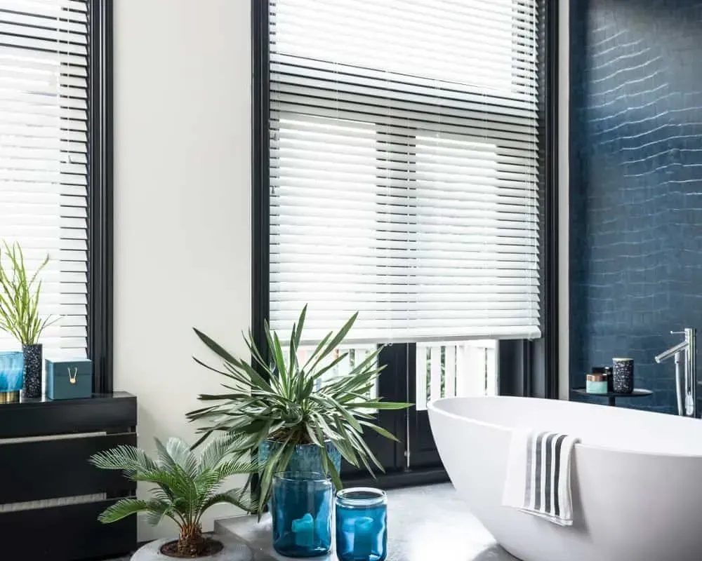 Badeværelse med hvidt badekar, planter, blå væg og persienner.