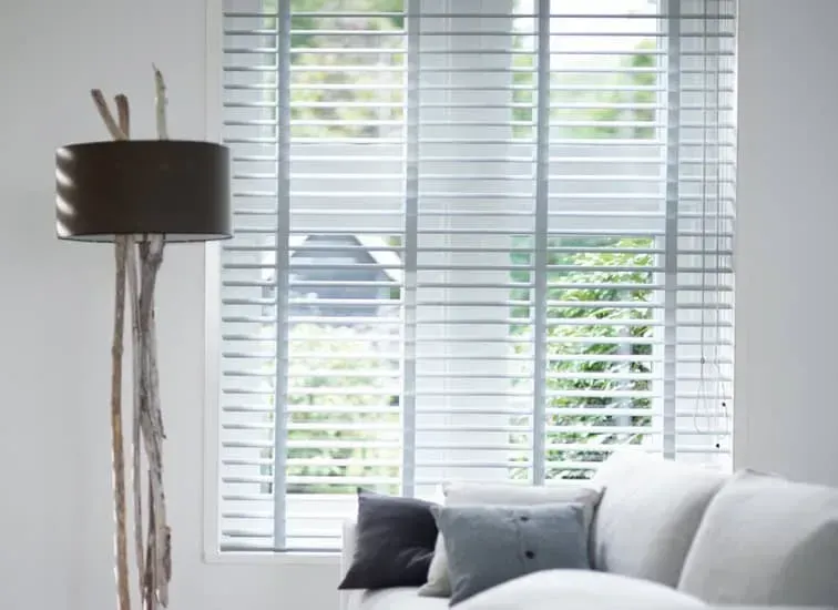 Stue med vindue, hvide persienner, en gulvlampe og en hvid sofa med puder.