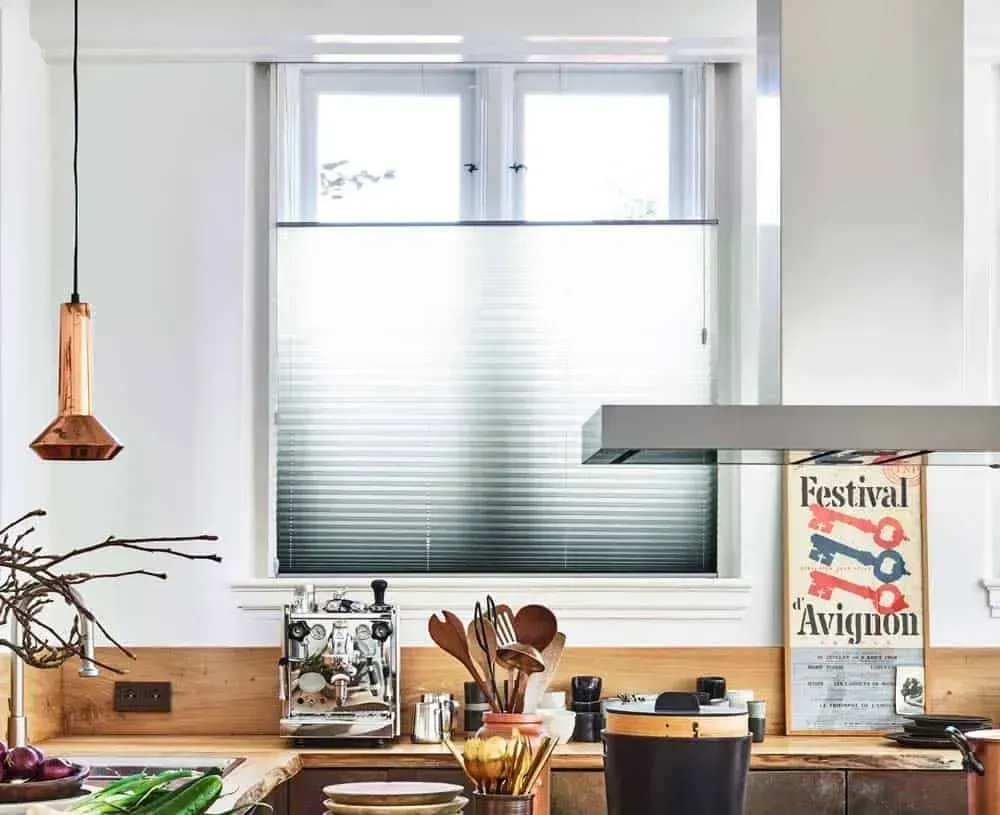 Køkken med vindue og persienner, bordplader i lyse træ, espressomaskine og kobberpendellampe.