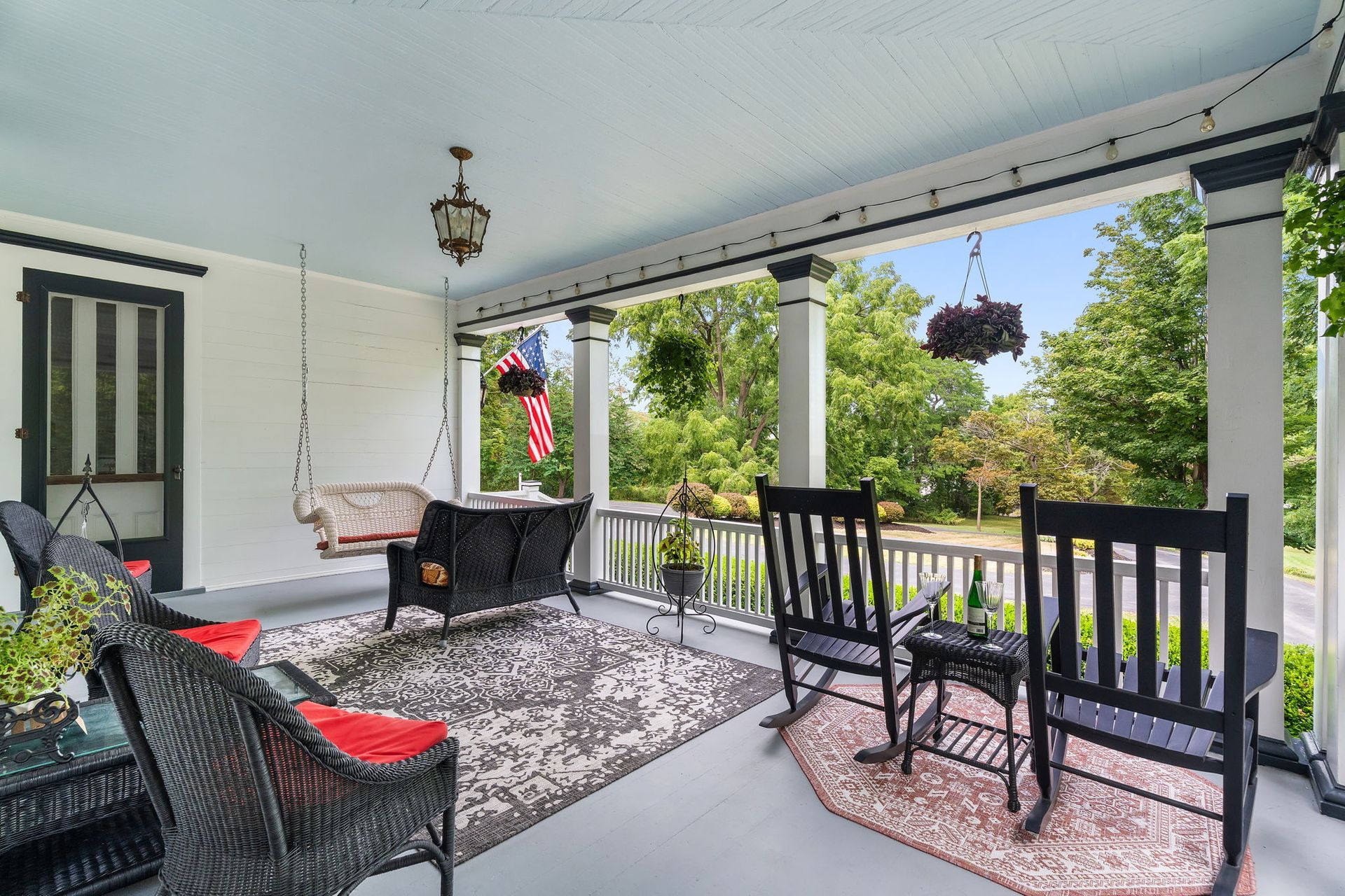 Covered front porch with black wicker chairs, a small table, hanging plants, and a view of green trees.
