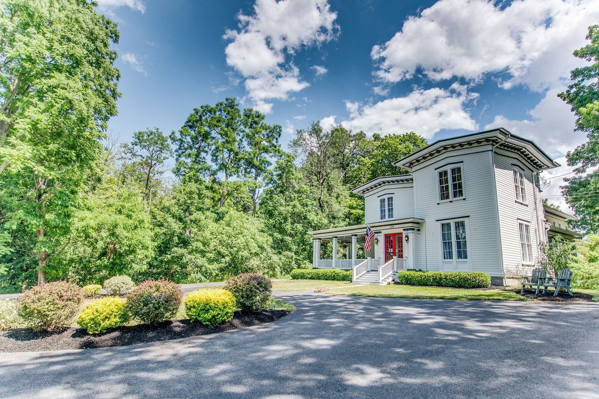 White two-story house with red door, framed by trees and shrubs under a blue sky