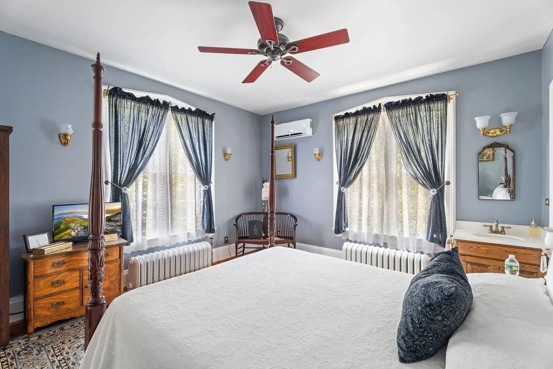 Bright bedroom with a white bed, blue curtains, red ceiling fan, and wooden furniture with mirrored nightstands