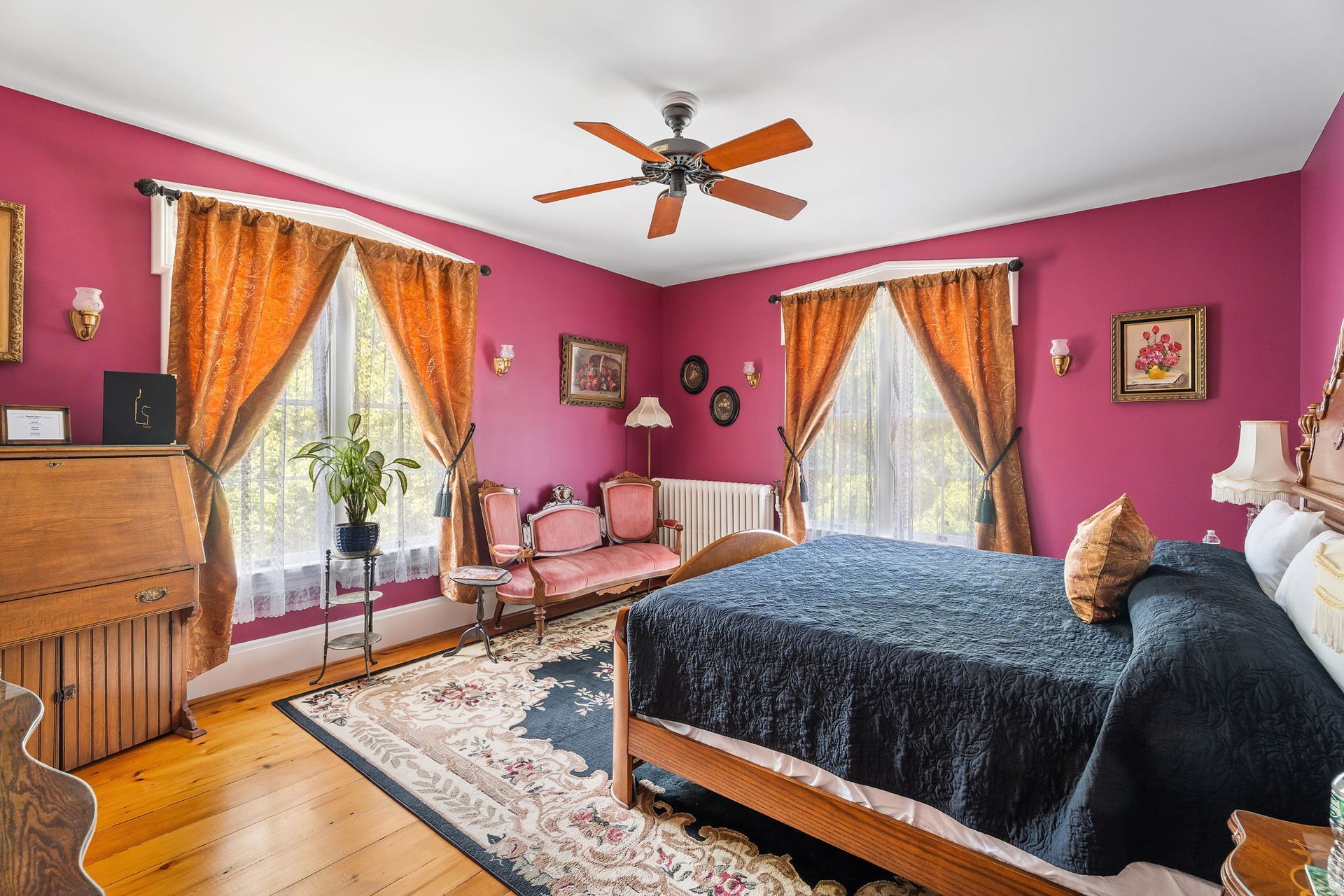 Bright pink bedroom with a bed, two chairs by windows, a dresser, and a ceiling fan.