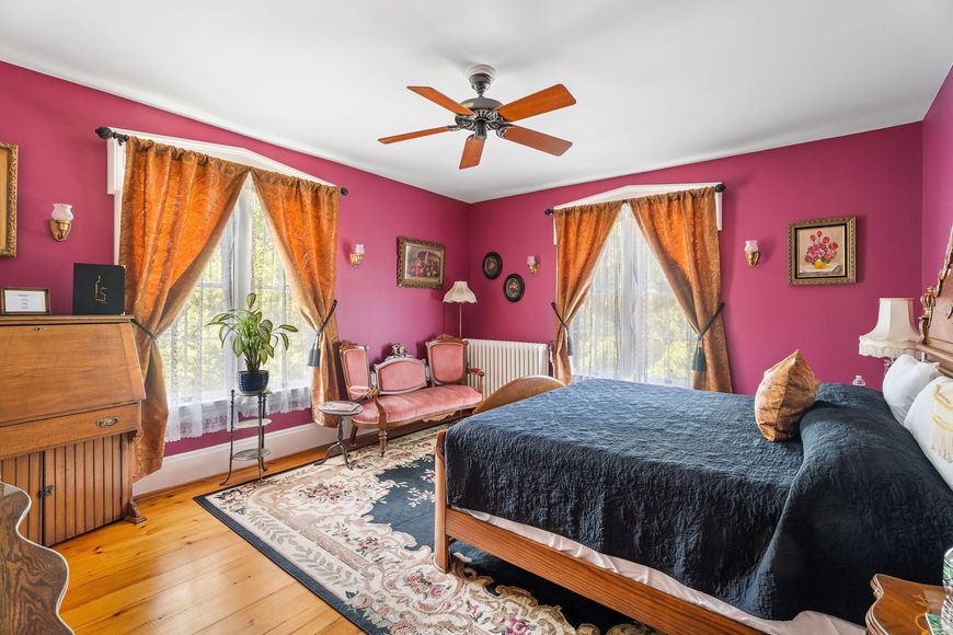 Bright pink bedroom with a bed, two chairs by windows, a dresser, and a ceiling fan.