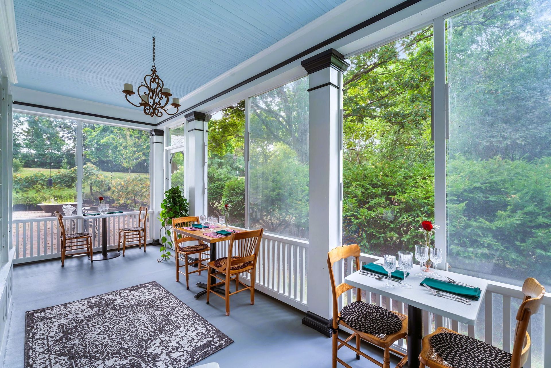 Bright sunroom with wicker tables and chairs, patterned rug, and large windows overlooking lush greenery