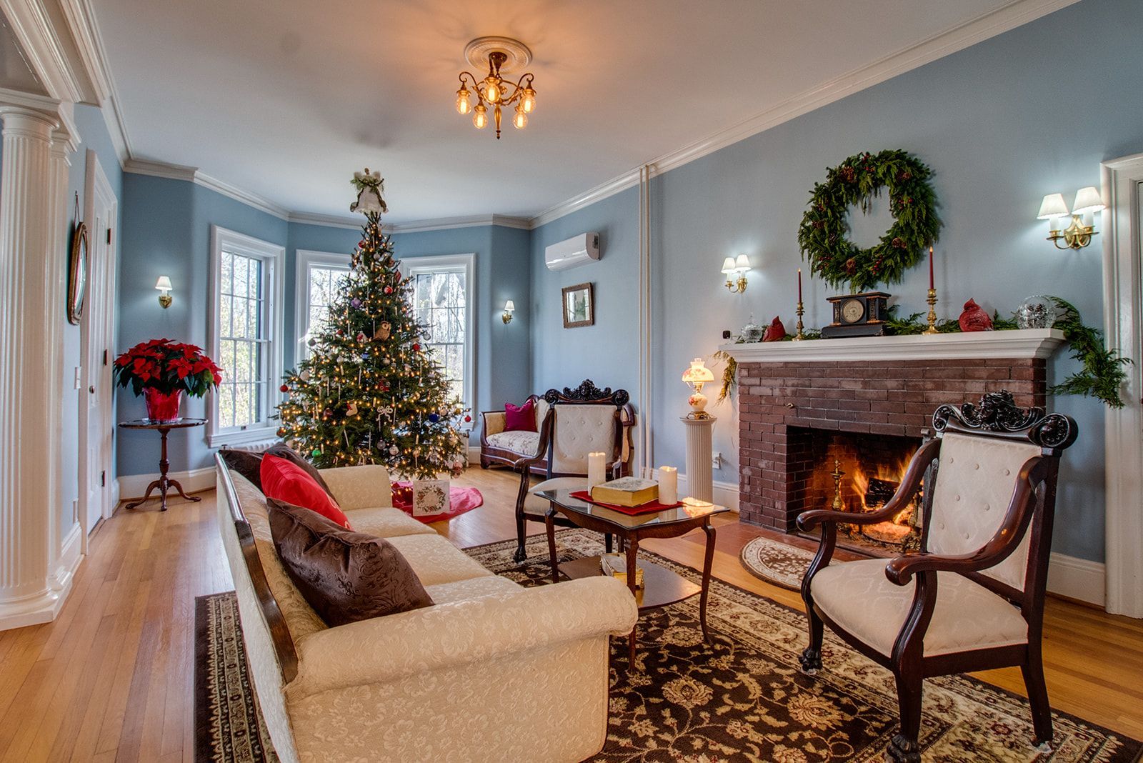 Cozy living room with lit Christmas tree, fireplace, wreath, and classic seating in soft blue tones