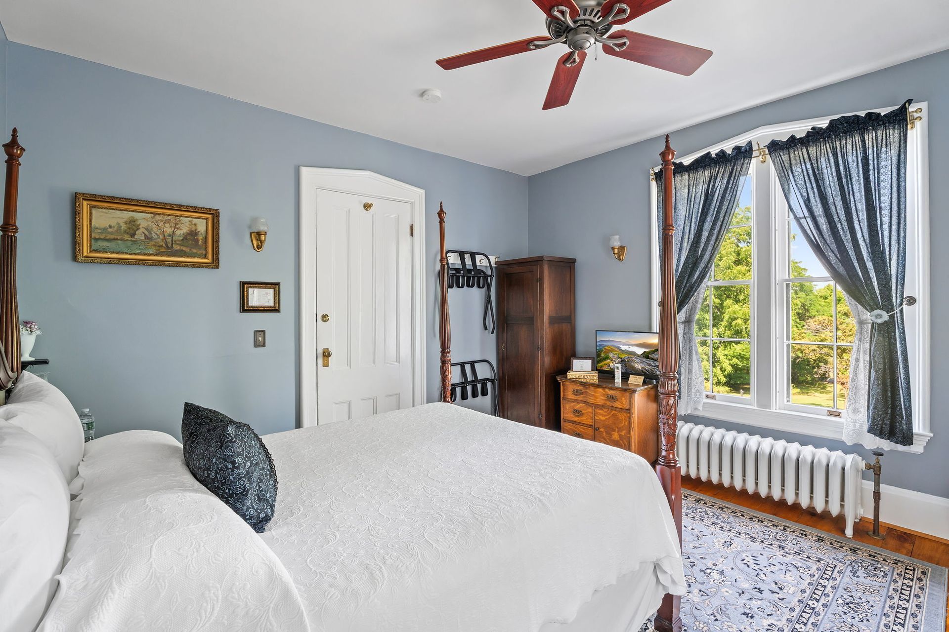 Cozy bedroom with white bed, blue walls, dresser, ceiling fan, and a bright window with curtains